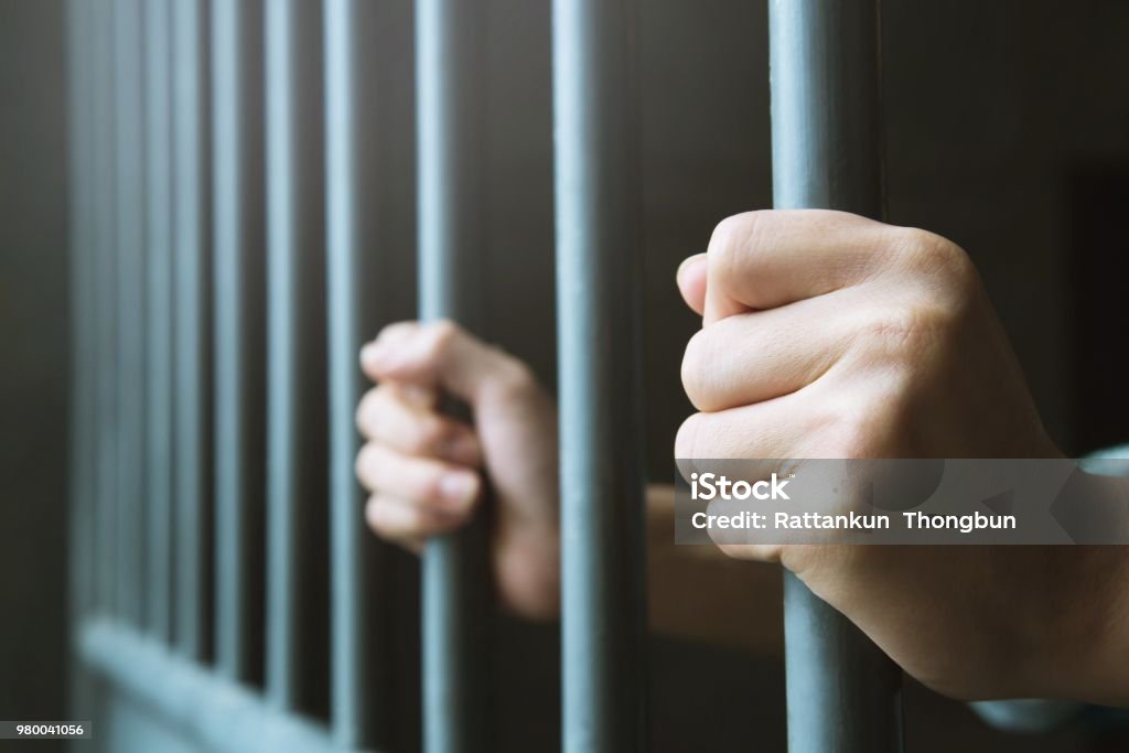 Човек во затвор со рацете одзади ги држи затворските решетки од челичен кафез.сторителот криминалец затворен во затвор.